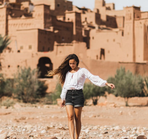 A young woman with long, wavy hair wears a white peasant blouse and black shorts, walking on a rocky, dusty path with traditional mud-brick buildings in the background.