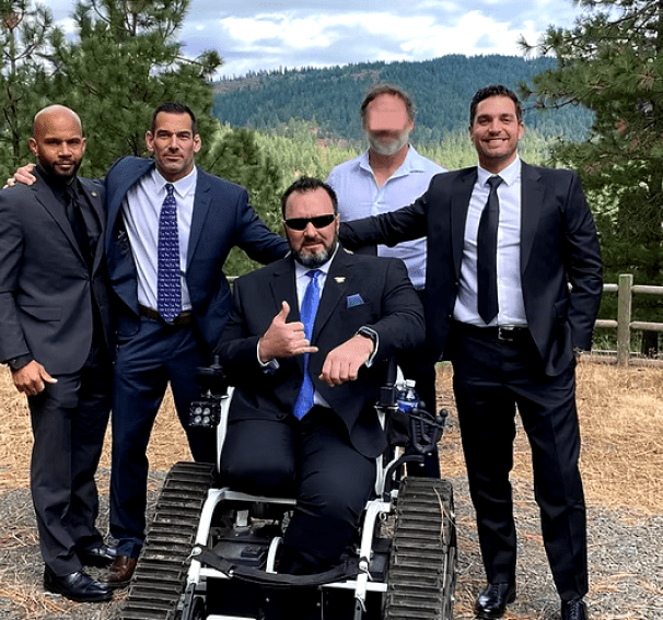 A group of five men, mostly dressed in suits, pose outdoors with a mountainous, forested background. One man is seated in an all-terrain wheelchair, giving a "shaka" sign. Another man's face is blurred.