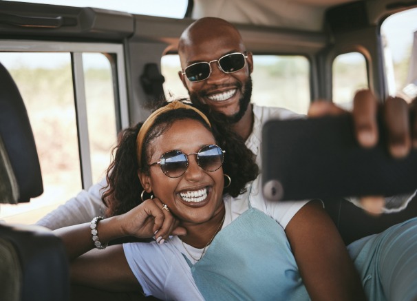 A happy couple in sunglasses smiles brightly while taking a selfie inside a car, likely on a road trip or safari.