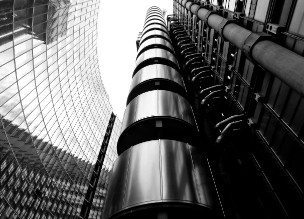 A low-angle, black and white architectural shot of two modern skyscrapers, one with a highly reflective glass facade and the other featuring prominent external pipes and cylindrical structures.