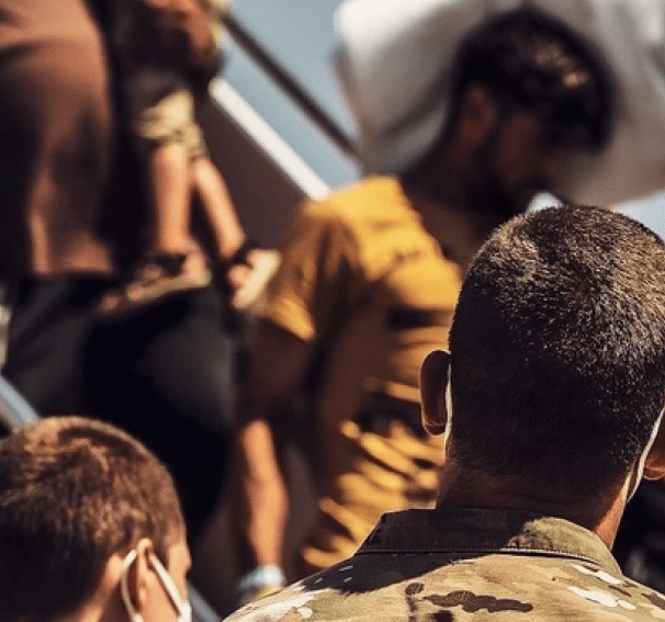 A blurred close-up from behind shows people boarding what appears to be a plane or large vehicle, with one person in the foreground wearing a camouflage shirt and another with a face mask.