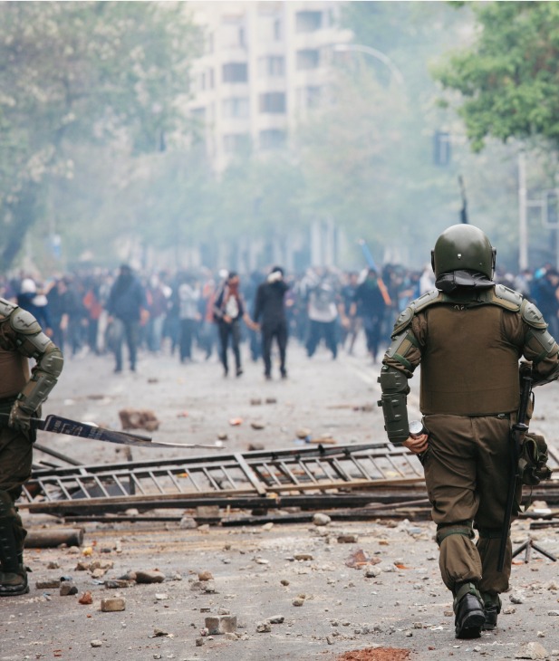 Two riot police officers face a large crowd of protesters on a debris-strewn street, with smoke or tear gas visible in the air.