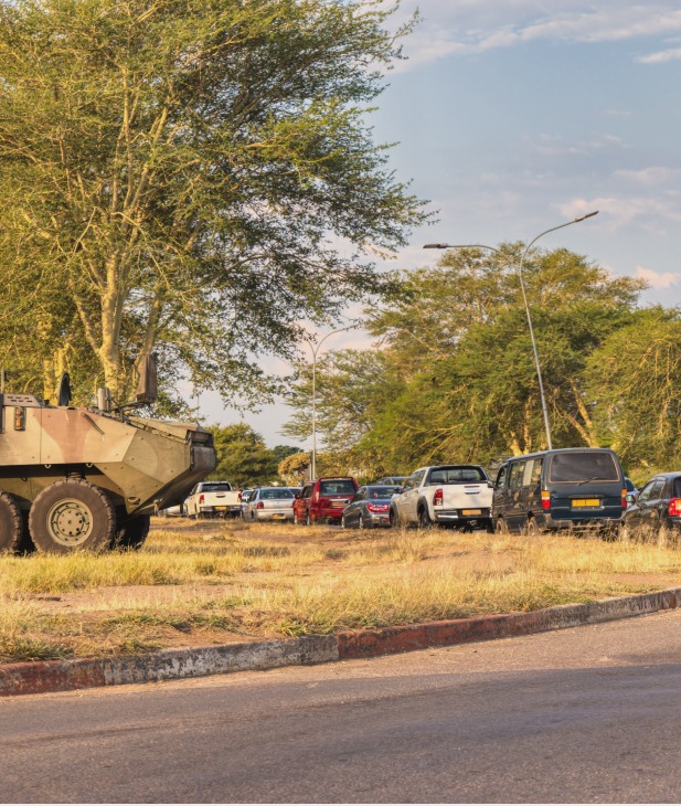 An armored military vehicle is parked on a grassy area next to a paved road, with several civilian cars parked behind it and trees under a partly cloudy sky.