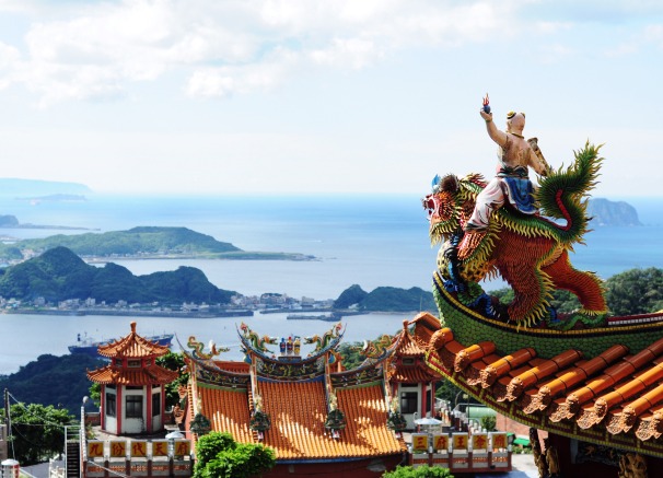 A vibrant, ornate dragon and figure statue adorns the roof of a traditional temple in the foreground, with a panoramic view of a bay, islands, and distant mountains under a blue sky in the background.