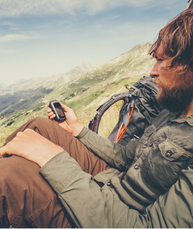 A male hiker with a beard, wearing a green shirt and brown pants, sits in a mountainous landscape and looks at a handheld GPS device. A backpack is beside him.